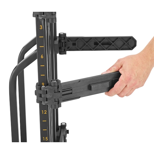 A close-up image of a DEWALT photography equipment stand, showing a person's hand adjusting a black arm attached to the stand. The stand features a vertical pole with yellow measurements marked from 3 to 15.