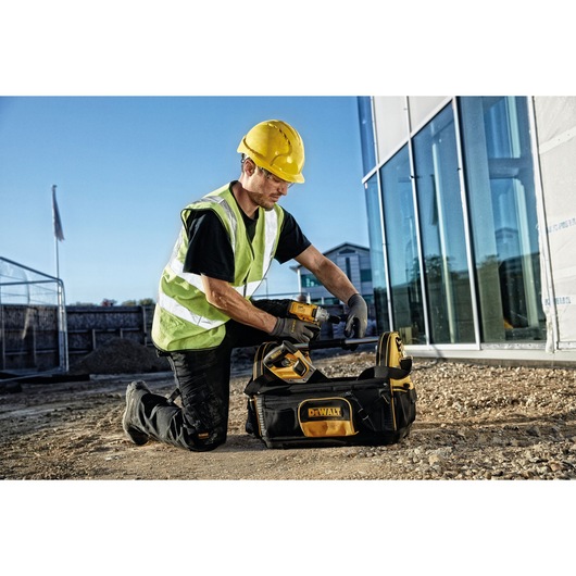 A construction worker wearing a yellow hard hat and reflective vest kneels outdoors next to a DEWALT tool bag, holding a power tool. The tool bag and power tool both display the DEWALT brand. The background shows a building with large glass windows.