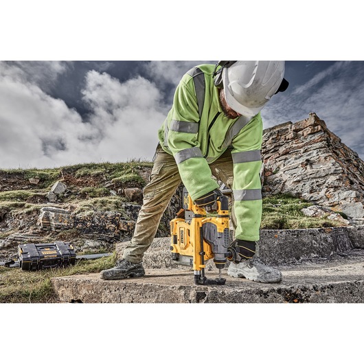 A worker wearing a hard hat and high-visibility jacket operates a yellow DEWALT rotary hammer drill on a concrete surface outdoors, with rocky terrain and a tool case visible in the background.
