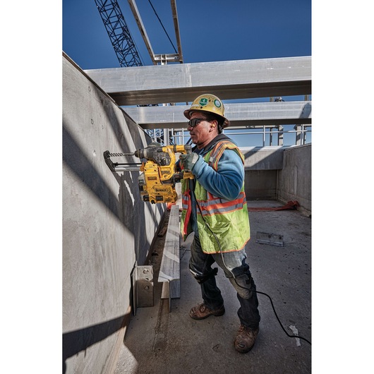 A construction worker wearing a safety vest and helmet is using a DEWALT D25333K rotary hammer drill to drill into a concrete wall at a construction site. The worker's face is blurred for privacy.
