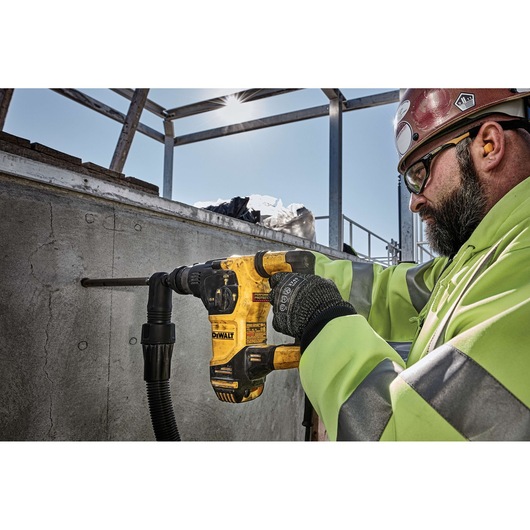 A person wearing safety gear is using a yellow DEWALT power drill to make a hole in a concrete wall at a construction site. The person's face is not visible clearly.