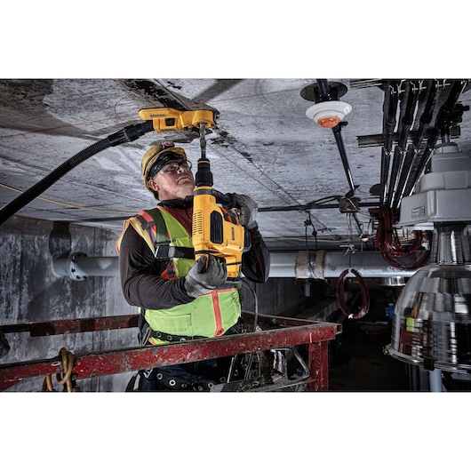 A construction worker wearing a safety vest and helmet is using a yellow DEWALT rotary hammer drill (SKU: D25481K) to drill into a concrete ceiling. The worker is standing on an elevated platform and the drill is attached to a dust extraction system for a clean work environment.