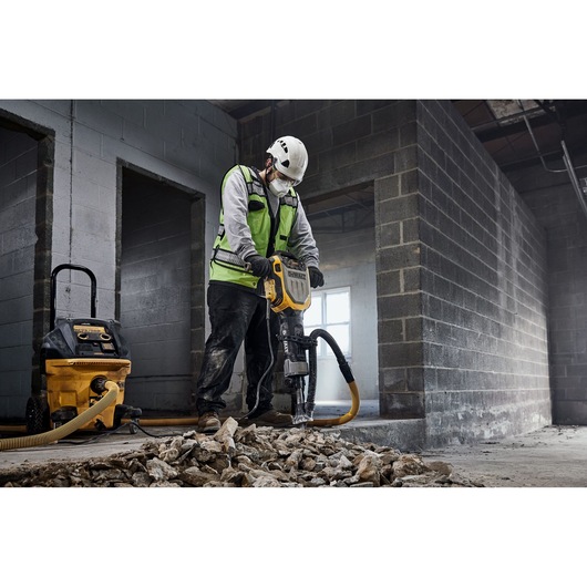 A construction worker wearing a safety vest and helmet is using a DEWALT jackhammer (model D25966) to break up concrete inside an unfinished building. There is a DEWALT dust extractor connected to the jackhammer, and broken concrete rubble is scattered on the floor.