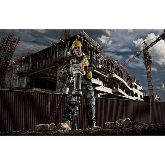 A construction worker wearing safety gear operates a DEWALT D25981 jackhammer on a construction site, with a large unfinished building and crane in the background.