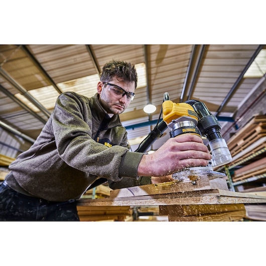 A person using a DEWALT D26200 wood router in a workshop, working on a wooden plank. The tool is connected to a dust extraction system. The person's face is blurred for privacy.