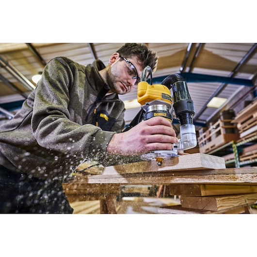 A person using a DEWALT D26200 wood router to shape a piece of wood in a workshop. Wood shavings are being produced as the router moves along the wood surface.