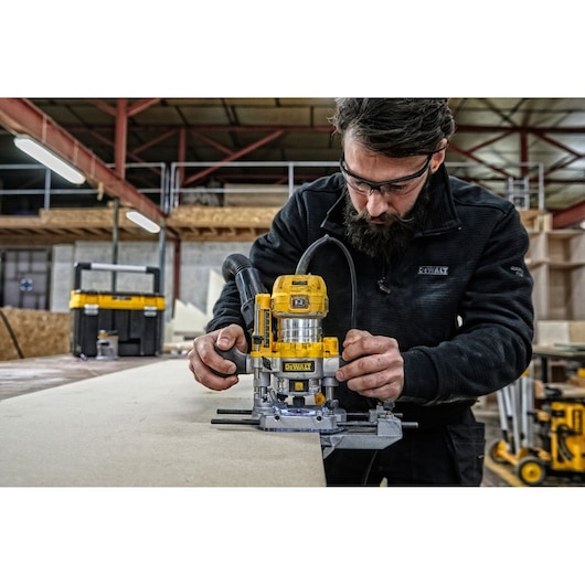 A person is using a DEWALT D26204K wood router on a workbench in a workshop. The router is yellow and black, and there is DEWALT equipment visible in the background. The person's face is blurred.