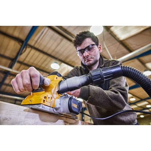 A person using a DEWALT D26500K electric planer with a connected dust extraction hose on a wooden plank inside a workshop. The user's face is blurred.