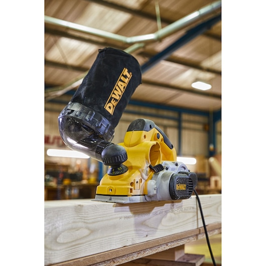 A DEWALT electric hand planer with dust bag attachment, placed on a wooden plank in a workshop environment.