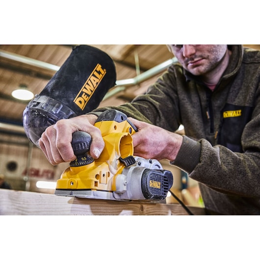 A person using a DEWALT electric planer (model D26500K) on a wooden plank in a workshop. The dust bag and DEWALT branding are clearly visible.