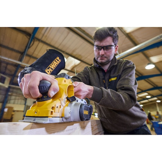 A person using a yellow DEWALT D26500K electric planer to smooth a wooden plank in a workshop. The tool has a dust bag attached and the person is wearing a DEWALT-branded sweater. The person's face is blurred for privacy.
