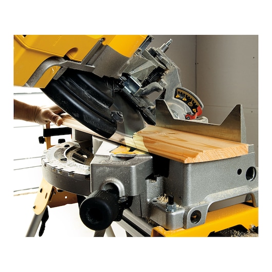 A DEWALT miter saw cutting a piece of wood, with a person's hand adjusting the material on the saw table. The saw has a metal base and a yellow upper body.