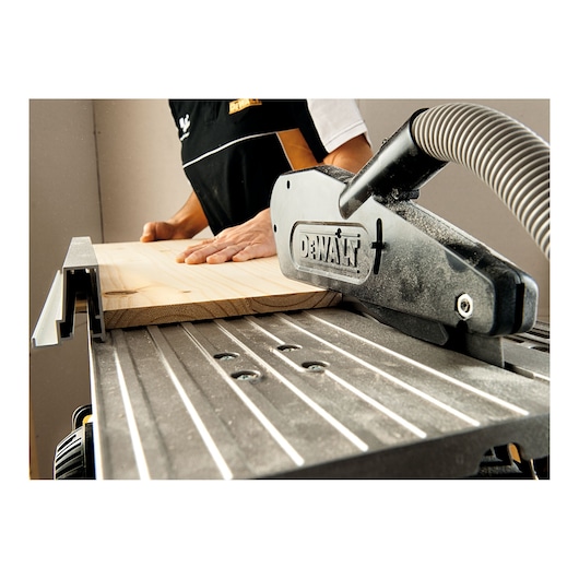 A DEWALT D27113 woodworking machine in use, with a person guiding a wooden plank through the saw. The machine features a dust extraction hose and safety guard.