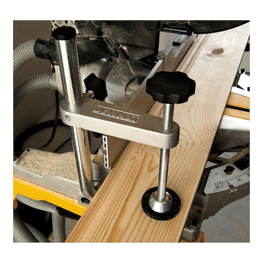 A close-up view of a DEWALT mitre saw with a wood board clamped securely using a metal clamp. The saw is set up for precise woodworking with adjustment knobs and a dust extraction hose visible in the background.