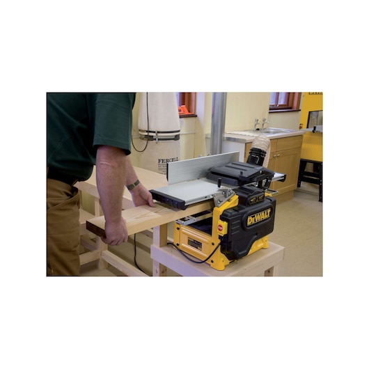 A person using a DEWALT wood planer machine (model D27300) to plane a piece of wood in a workshop setting.