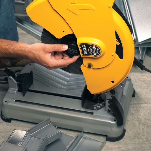 A close-up of a person adjusting a DEWALT D28715 yellow chop saw, used for cutting metal pieces. Several metal angles are visible around the saw base on a workshop floor.