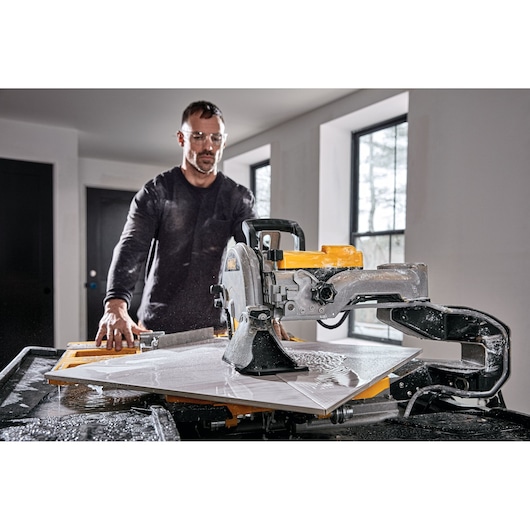 A person operates a DEWALT D36000 wet tile saw on a worktable, cutting a large white tile inside a modern room. The saw is actively spraying water to cool the blade during the cutting process.