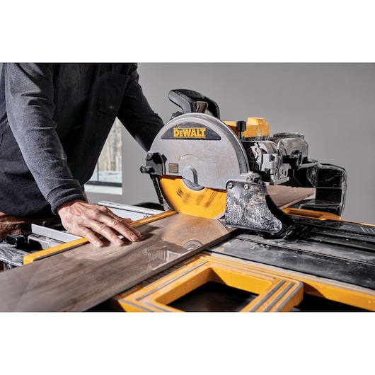 A person is using a DEWALT D36000 wet tile saw to cut a piece of tile. The saw is yellow and black, and the worker is guiding the tile on the cutting platform.