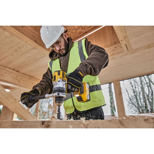 A person wearing a high-visibility vest and hard hat uses a DEWALT cordless power drill to bore a hole into a wooden beam on a construction site. Wood shavings are visible as the drill operates.