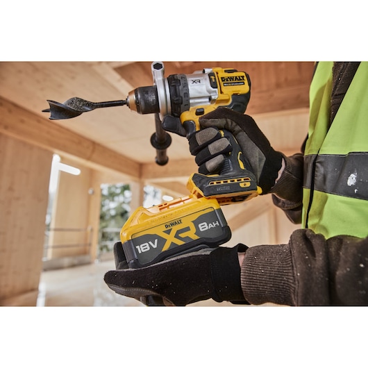 A close-up of a DEWALT power drill in use, showing a gloved hand holding the drill. The drill is equipped with an 18V XR 8Ah battery, and the scene appears to be in a wooden construction environment.