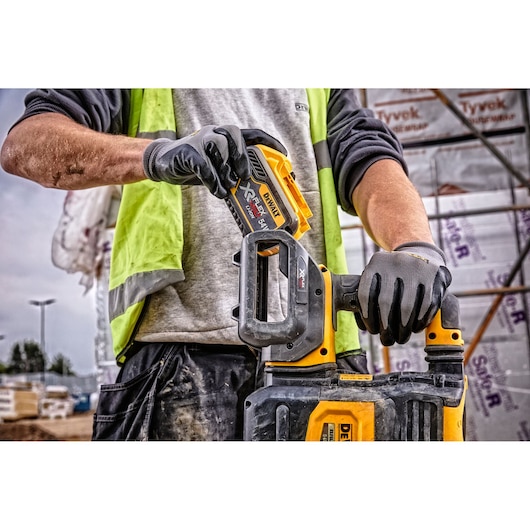 A person wearing work gloves and a high-visibility vest is inserting a DEWALT XR FLEXVOLT battery into a yellow and black power tool at a construction site.