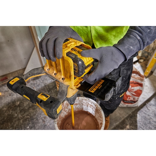 A person wearing gloves uses a DEWALT cordless power tool with a DCB548 battery to mix material in a bucket at a construction site.