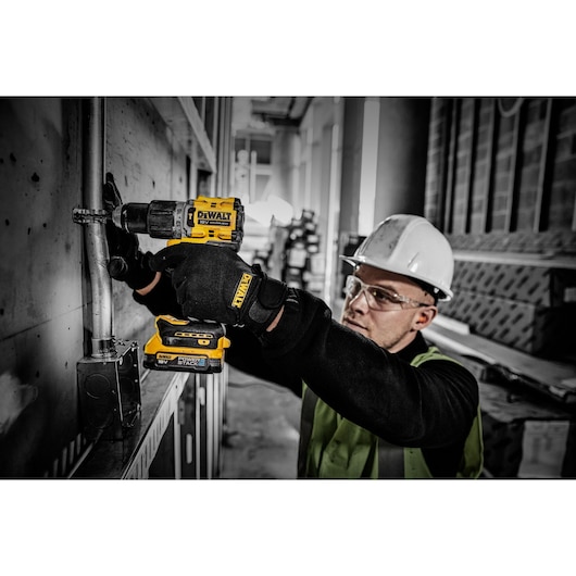 A person wearing a hard hat and safety vest is using a DEWALT cordless drill with a DCBP034 Power Stack battery to work on a metal pipe attached to a wall in a construction environment.
