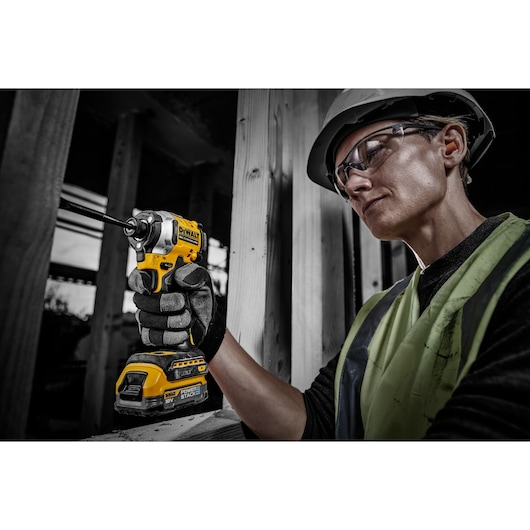 A person wearing work gloves and a safety vest holds a DEWALT power tool with a POWERSTACK 18V battery. The person's face is blurred and they appear to be working at a construction site.