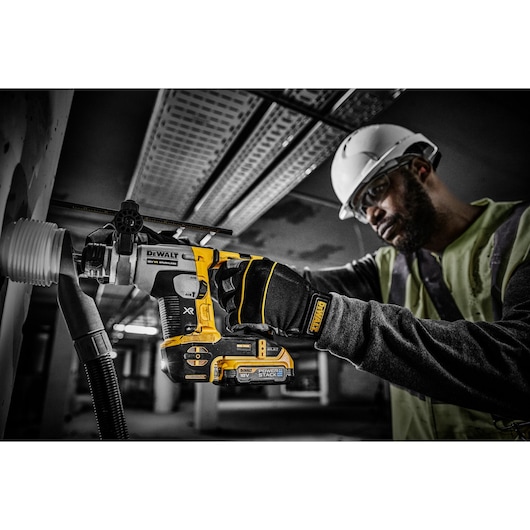 A person wearing safety gear uses a DEWALT cordless power drill with a DCBP034 POWERSTACK 18V battery to drill into a wall in an industrial environment.