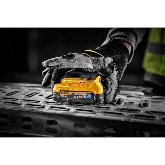 A gloved hand holding a DEWALT 18V POWERSTACK lithium-ion battery, placed on a metallic surface in an industrial setting.