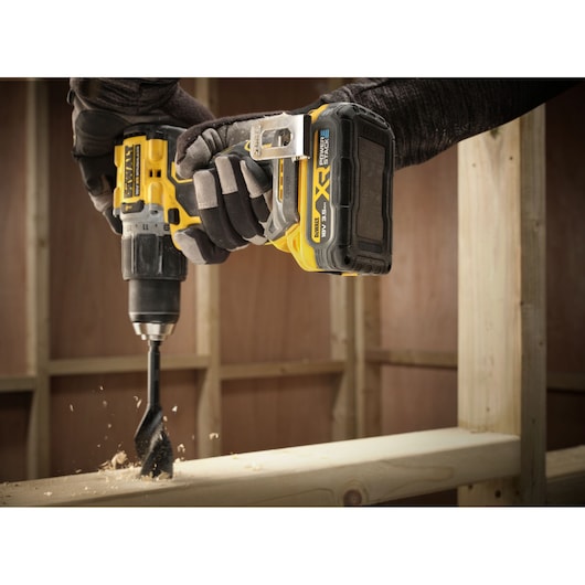 A close-up of a person using a yellow and black DEWALT cordless drill with a DCBP318 XR POWERSTACK 18V battery, drilling through a piece of wood, with wood shavings flying.
