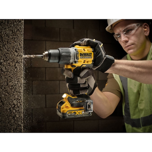 A gloved person using a DEWALT cordless power drill to bore into a rough concrete wall, with dust particles visible. The drill model shown is an 18V XR brushless device.