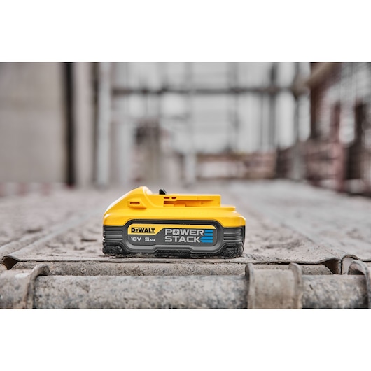 A DEWALT POWERSTACK 18V 5Ah battery lies on a construction site surface, with blurred construction materials in the background.