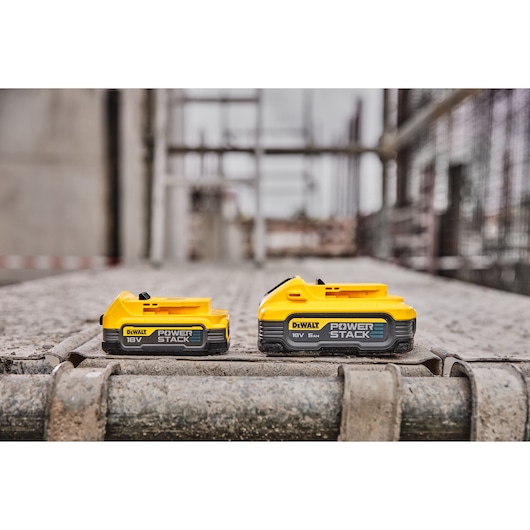 Two DEWALT POWERSTACK 18V batteries are shown side by side on a construction site, with a yellow and black color design. The batteries are placed on a metal surface with a blurred industrial background.