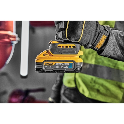 A close-up of a gloved hand holding a DEWALT POWERSTACK 18V 5AH battery pack. The background is blurred, highlighting a high-visibility vest and work environment.