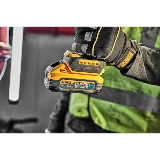 A person wearing a glove holds a DEWALT POWERSTACK 18V 5Ah lithium-ion battery, designed for power tools. The battery is yellow and black with branding visible.