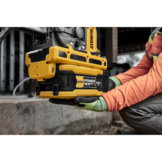 Close-up of a worker wearing gloves and an orange shirt holding a DEWALT POWER SHIFT DCBPS0554 battery pack attached to a DEWALT power tool at a construction site.