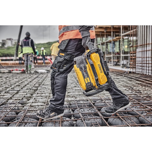 A person wearing work clothes and safety gloves is carrying a yellow DEWALT portable power station (SKU: DCBPS0554) across a construction site, surrounded by metal reinforcements and other workers in the background.