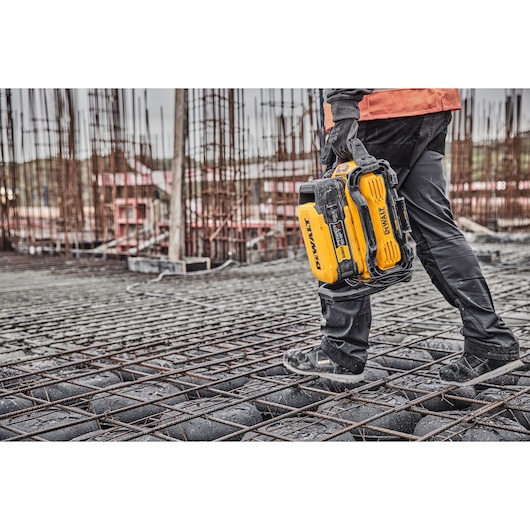 A worker wearing black pants and gloves carries a yellow DEWALT portable power station on a construction site with metal rebar and concrete blocks.