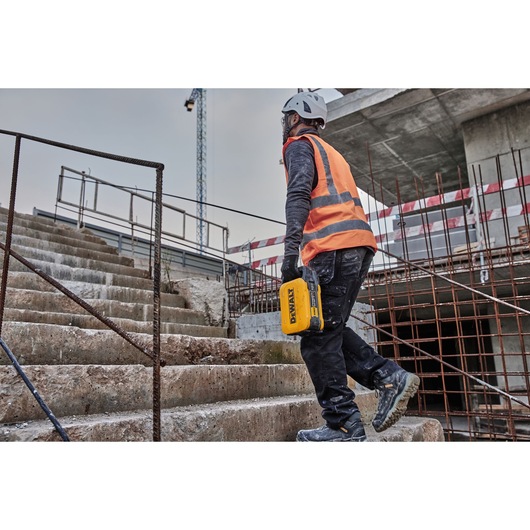 A person wearing a safety helmet and orange high-visibility vest is walking up concrete stairs at a construction site, holding a yellow DEWALT device.