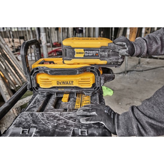 A DEWALT Power Shift 554 Wh battery being inserted into a DEWALT device by a person wearing work gloves, with construction materials visible in the background.