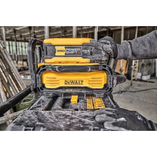 A close-up of a DEWALT POWER SHIFT 554 WH battery being placed onto a DEWALT charging station by a person wearing work gloves, set in an industrial construction environment.