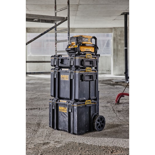 A stack of DEWALT ToughSystem tool storage boxes on wheels, shown on a construction site floor. The boxes are black with yellow accents and have handles for easy transport.