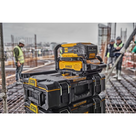 A DEWALT Power Shift DCBPS0554 battery sits on top of stacked DEWALT ToughSystem toolboxes at a construction site. Several workers in safety vests are visible in the background, with metal rods and building materials around.