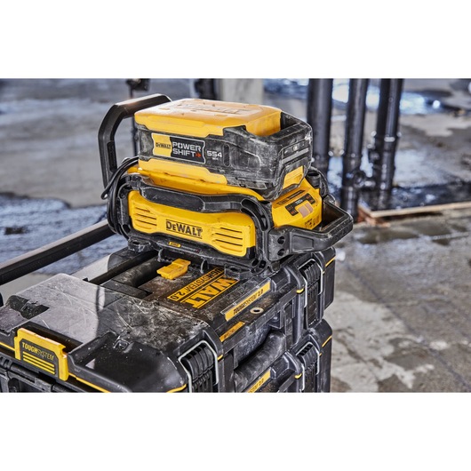 A DEWALT Power Shift battery (model DCBPS0554) is connected to a yellow and black DEWALT charger, placed on top of a stack of DEWALT ToughSystem toolboxes at a construction site with concrete floor and pipes in the background.