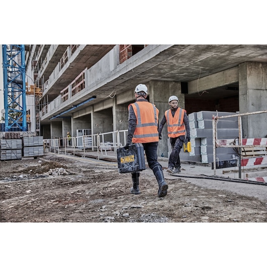Two construction workers wearing orange high-visibility vests and white helmets are walking on a building site. One is carrying a black DEWALT case, and the other holds a yellow tool. The site is unfinished with concrete structures and scattered building materials.