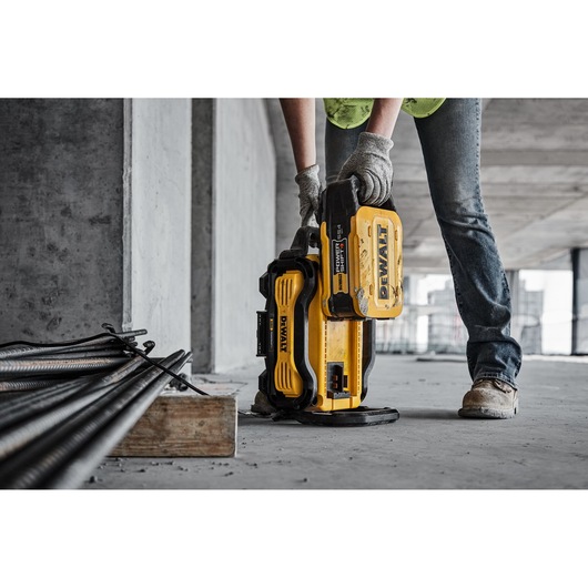 A person wearing gloves and work boots uses a DEWALT portable battery charger on a construction site, with metal rods and concrete surfaces visible.
