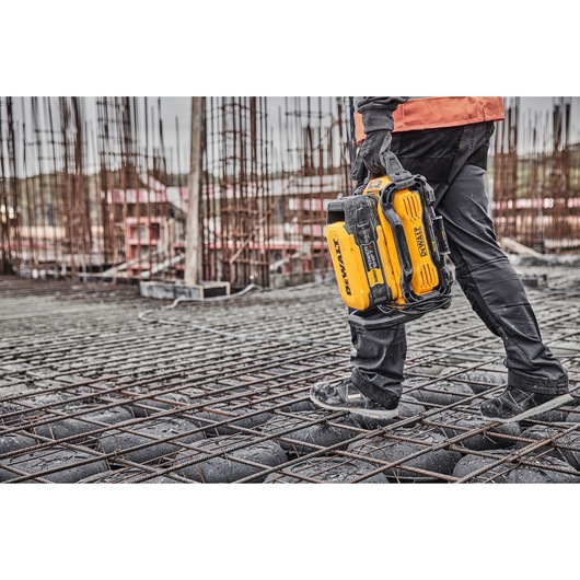 A person wearing construction gear is walking on a rebar grid at a construction site while carrying a yellow DEWALT portable power station.