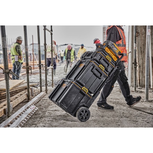A stack of DEWALT black and yellow tool storage boxes on wheels being transported by a person in an orange safety vest at a construction site, with several workers and building materials visible in the background.