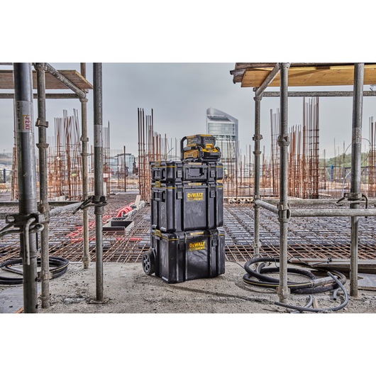 A stack of DEWALT black toolboxes, including a wheeled unit and two additional cases, is shown on a construction site with scaffolding and metal rebar in the background.